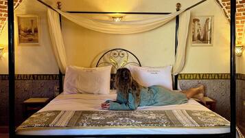 Una cama con dosel de madera con un edredón y almohadas blancos, estructura metálica negra y pared de ladrillo al fondo.