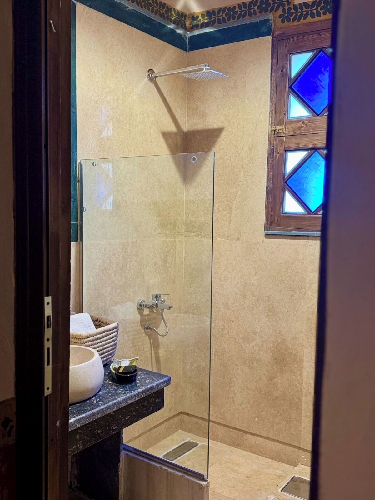 A glass-enclosed shower with a handheld showerhead, a small sink with a soap dish, and a decorative window with blue-tinted glass.