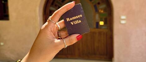 A hand holding a card with "Ramses Villa" written on it, in front of a building with an arched doorway.
