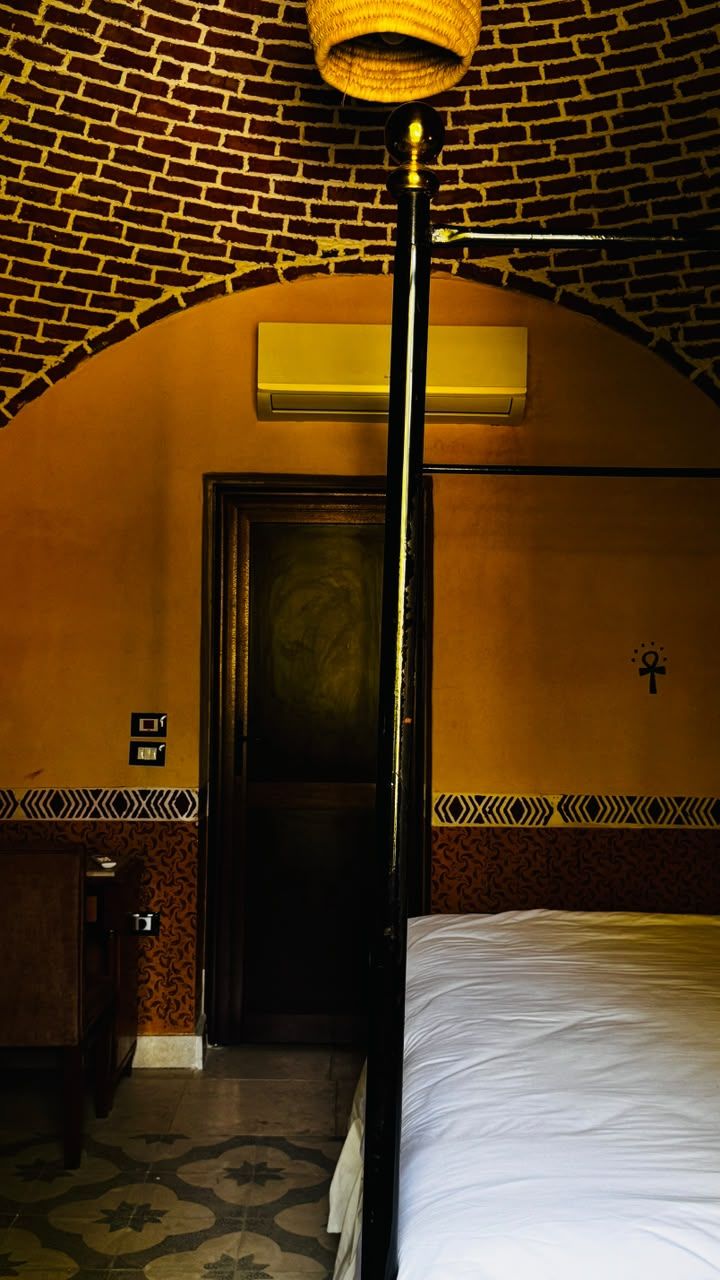 A hotel room with a brick-patterned ceiling, a bed with white linens, a door with a dark frame, and a patterned floor.