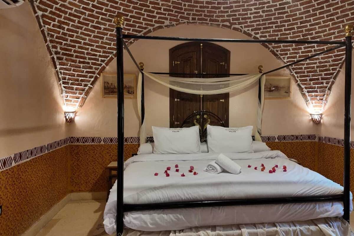 A four-poster bed with white linens and rose petals, set against a brick wall with patterned trim.