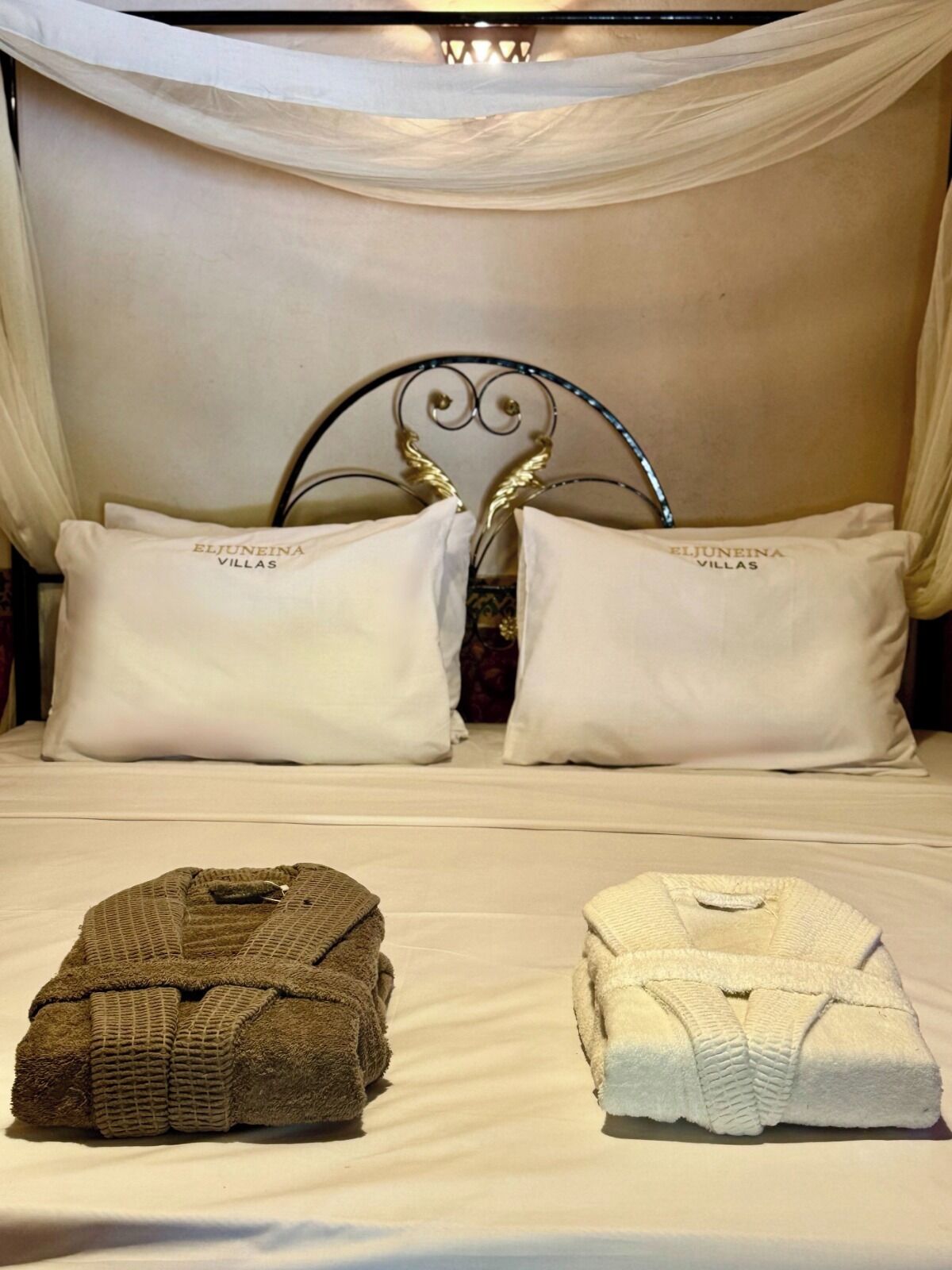 Ein Bett mit weißer Bettwäsche, zwei Kissen mit dem Aufdruck 'ELJUNEINA VILLAS', ein Metall-Kopfstück und zwei gefaltete Handtücher.