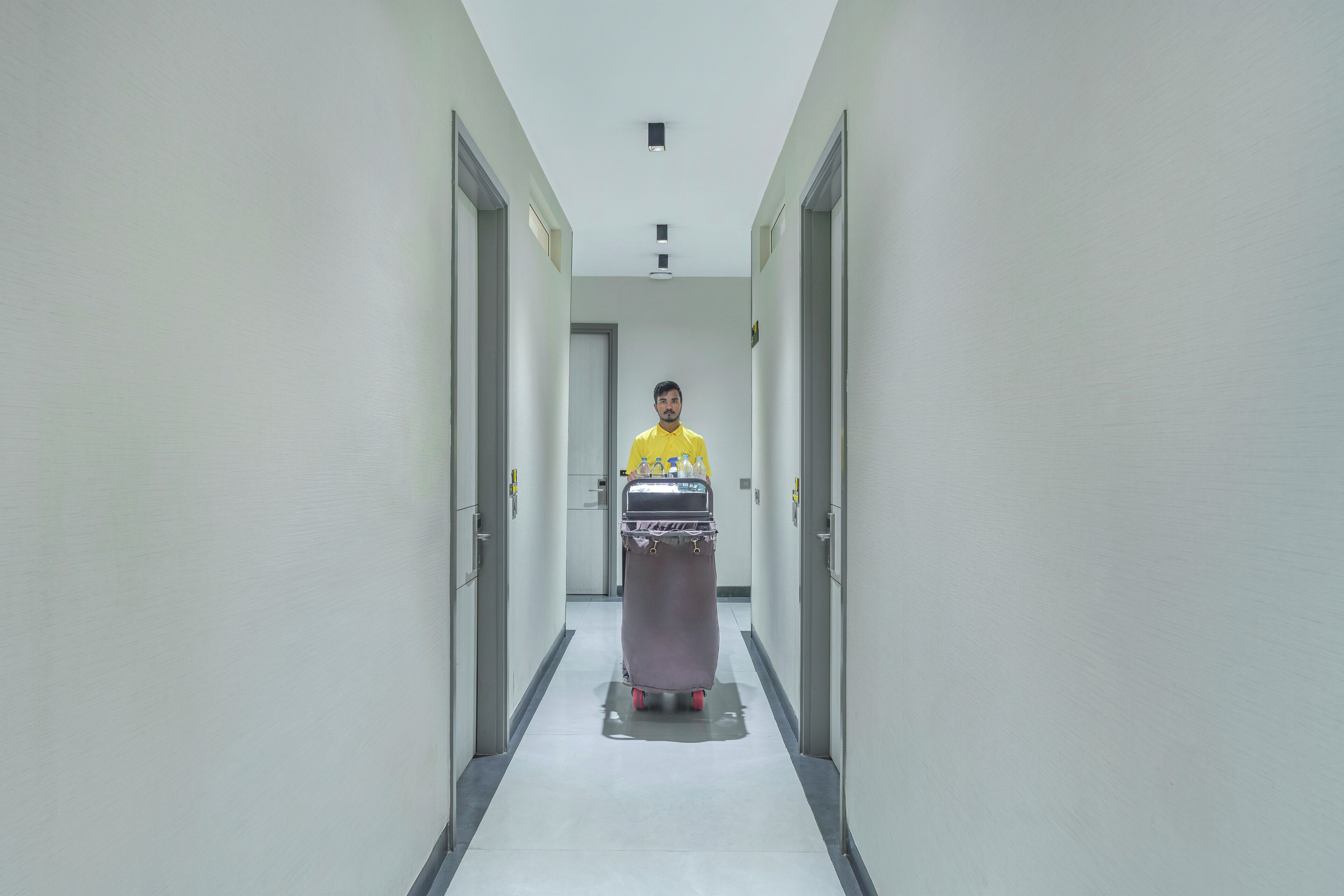 A person in a yellow shirt pushing a cart down a narrow, well-lit hallway with doors on either side.