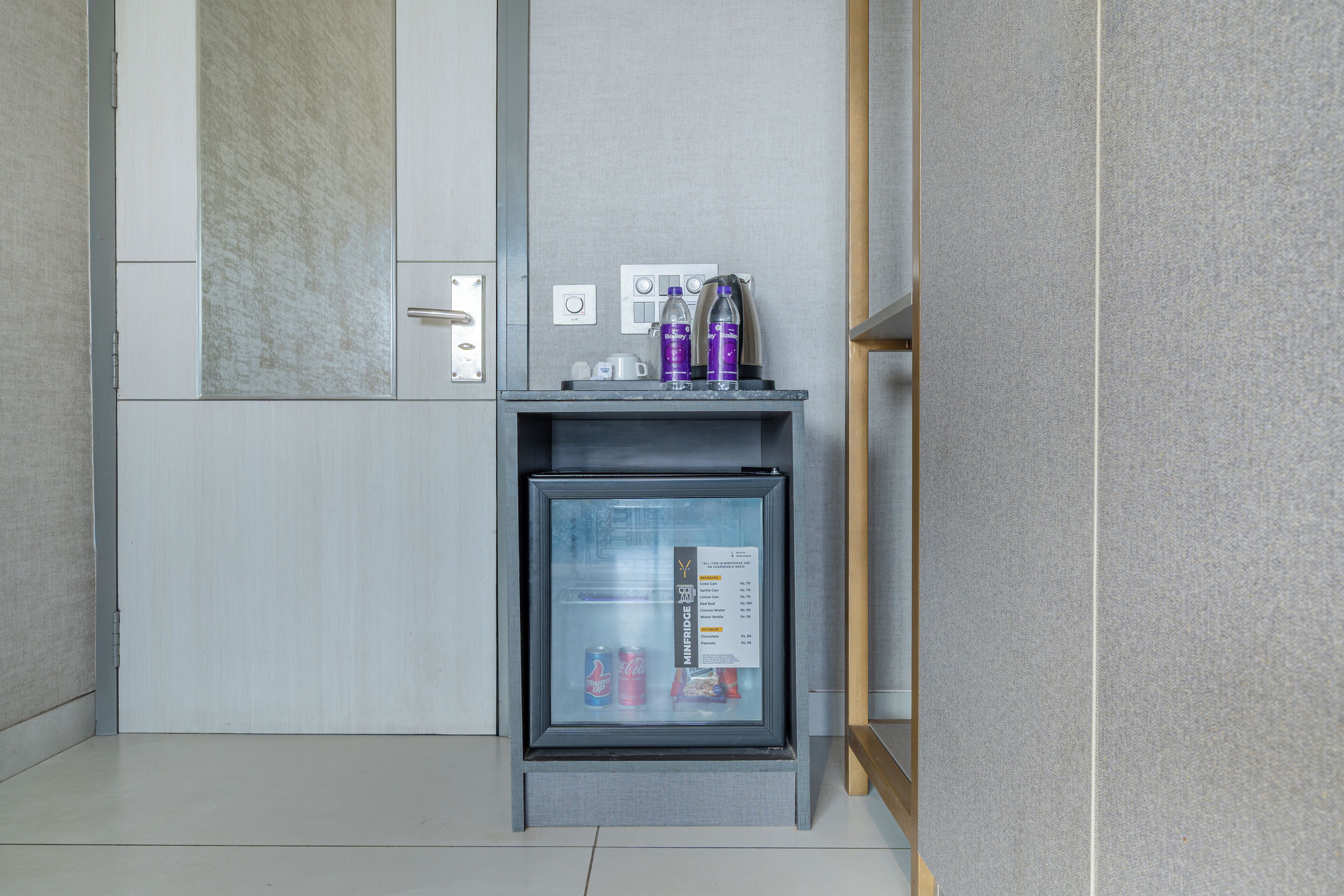 A minibar with a glass door, containing a water bottle, a can, and a small box.