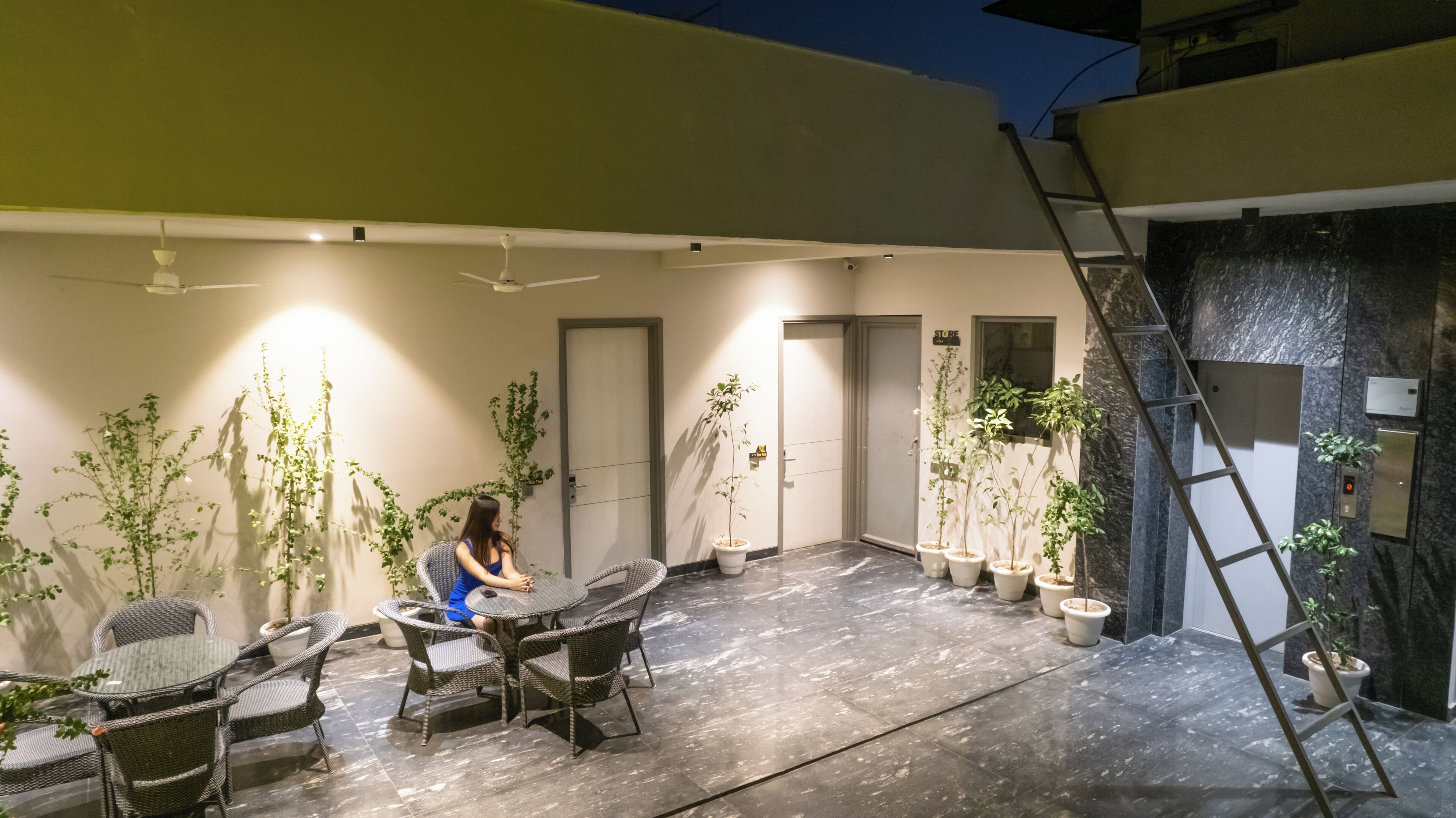 A woman sitting at a table in a well-lit outdoor area with potted plants and a tiled floor.