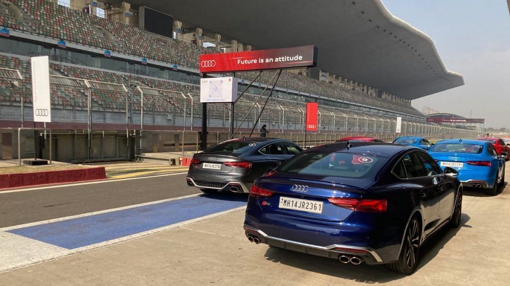 A parking area with multiple cars, including a blue Audi and a black Audi, in front of a stadium with empty stands and a large advertisement sign.
