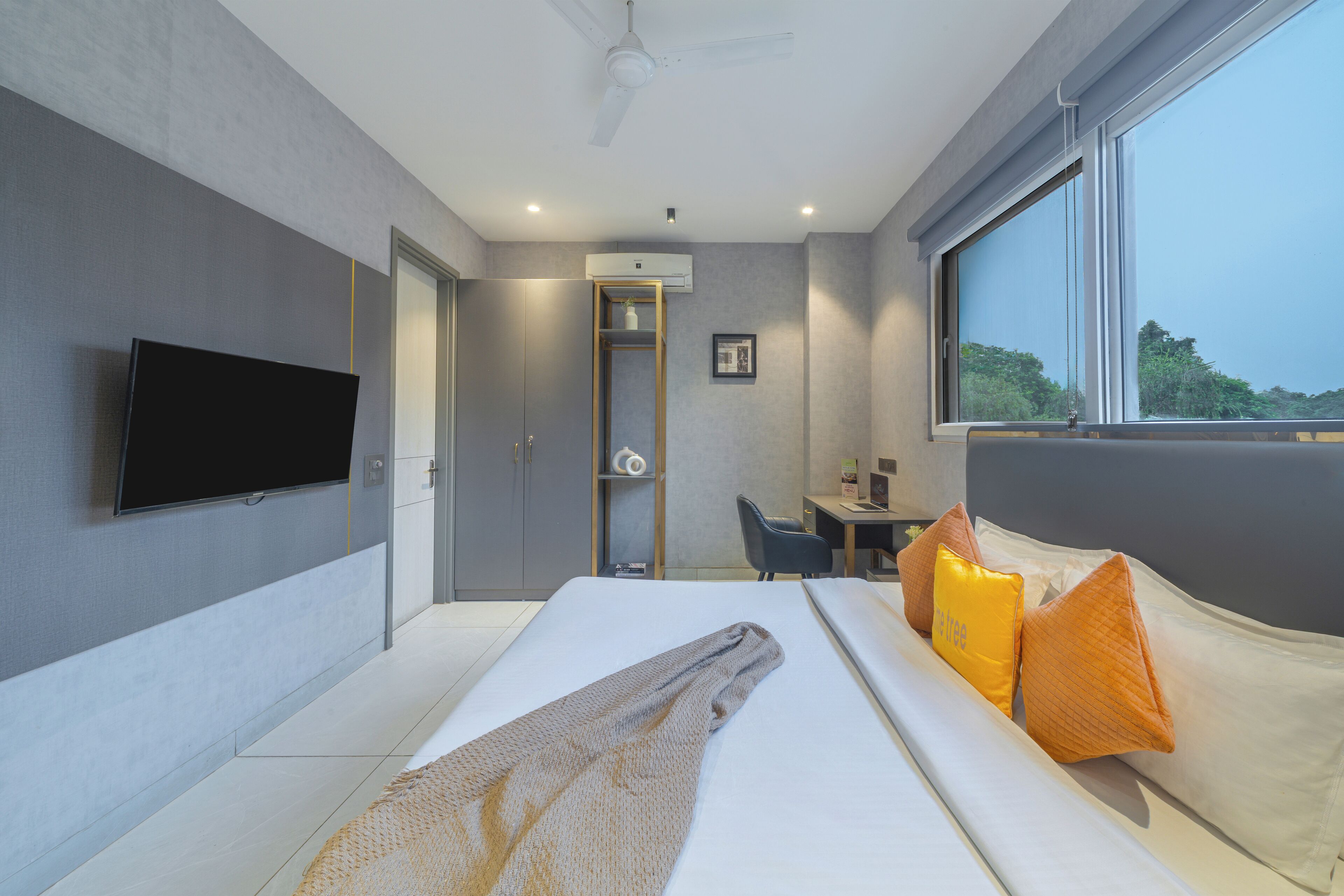 Une chambre d’hôtel moderne dotée d’une télévision à écran plat, d’un lit avec des oreillers blancs et orange, d’un bureau avec une chaise et d’une grande fenêtre donnant sur un paysage verdoyant.
