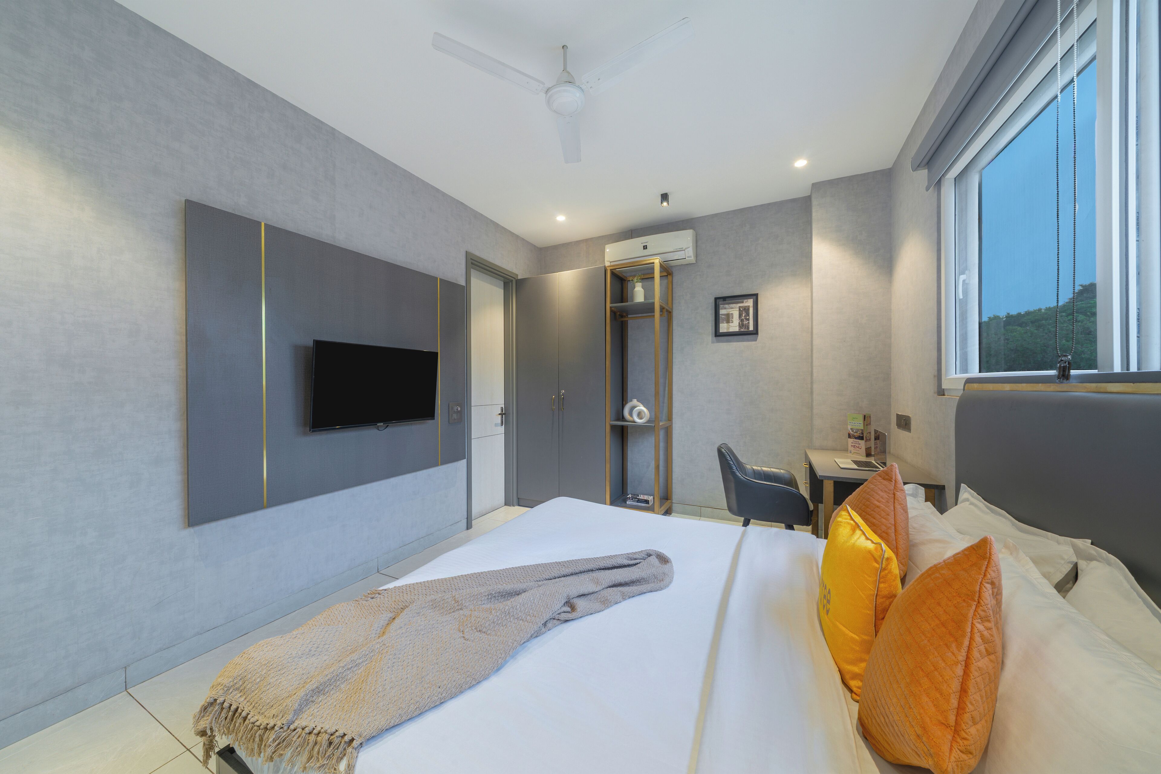 A modern hotel room with a bed, a flat-screen TV mounted on the wall, a desk with a chair, and a large window.