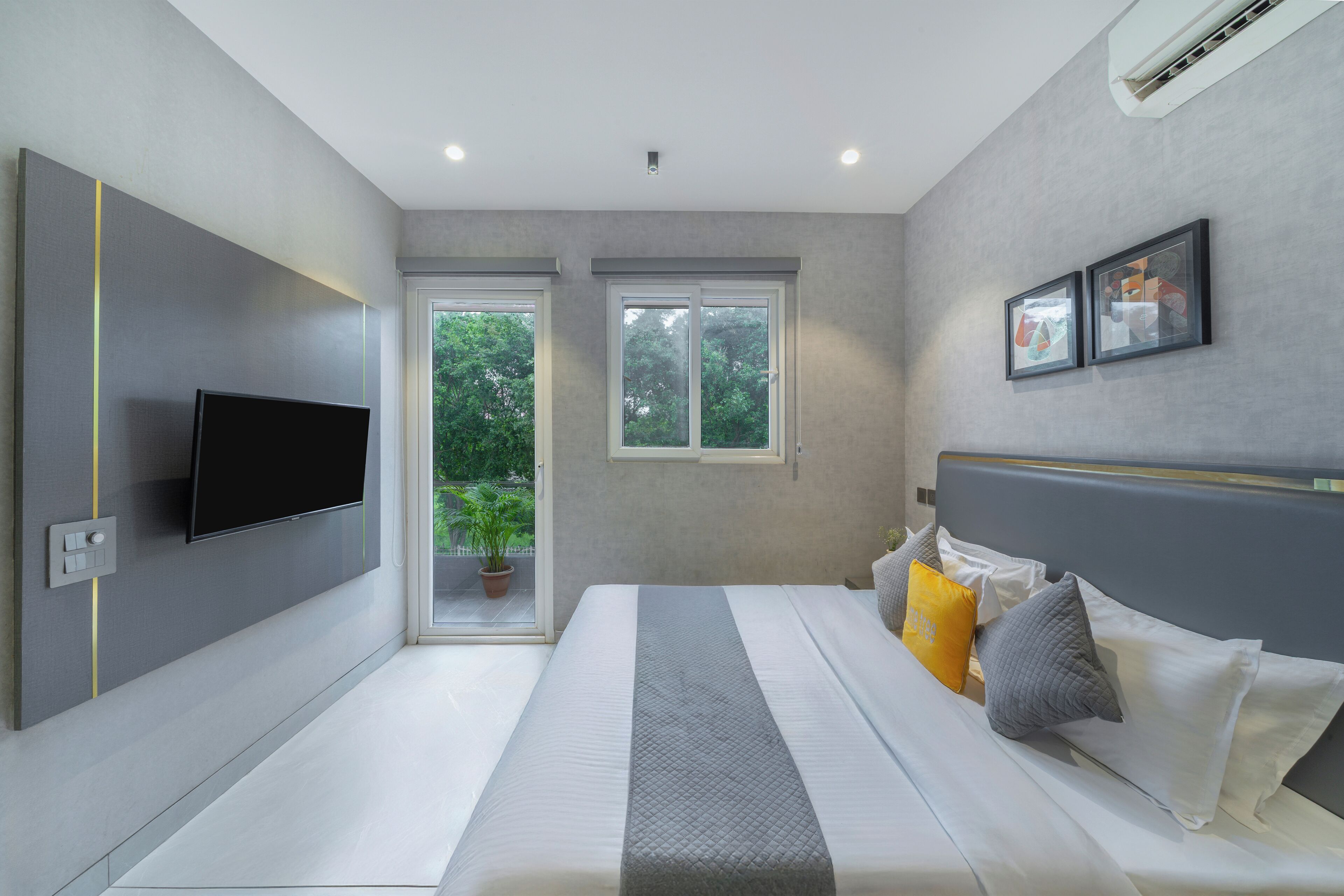 A modern bedroom with a large bed, a flat-screen TV mounted on the wall, and a window providing a view of greenery outside.