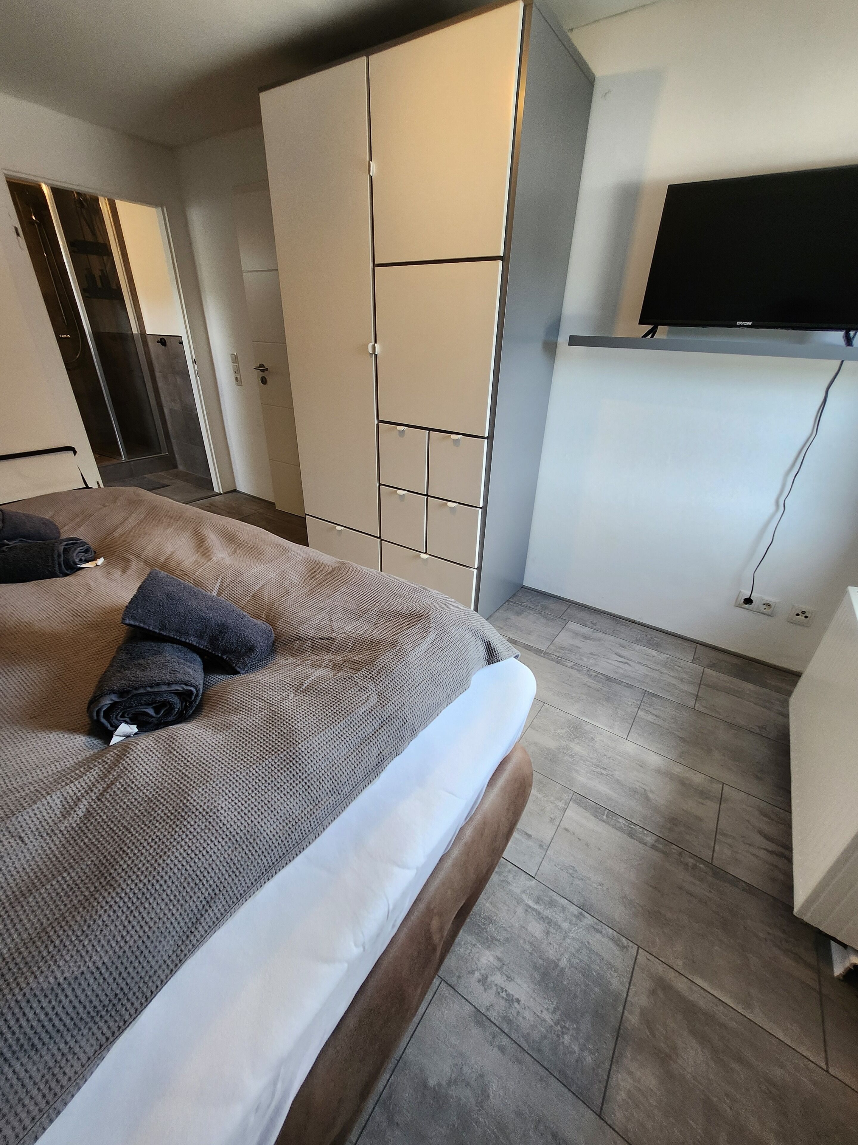 A modern bedroom with a bed, a wardrobe, a TV, and a shower area.