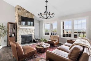 Un salón con muebles de cuero, chimenea de piedra, televisor de pantalla plana y ventanas amplias.