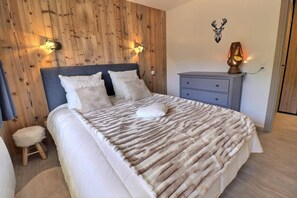 A bedroom with a wooden wall, a bed with a white blanket, two wall-mounted lights, a nightstand with a lamp, and a deer head decoration.