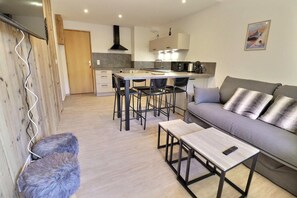 A modern living space with a grey sofa, a wooden bench, a dining area with chairs, a kitchen with a stove, and a wooden wall.