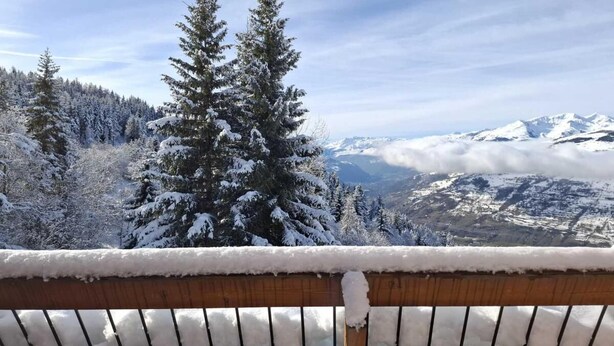 Une terrasse en bois recouverte de neige, offrant une vue sur un paysage montagneux enneigé et parsemé d’arbres à feuilles persistantes.