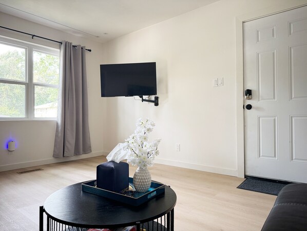 Una sala moderna con un televisor de pantalla plana montado en la pared, una mesa de centro redonda con un jarrón de flores blancas, y una puerta con felpudo.
