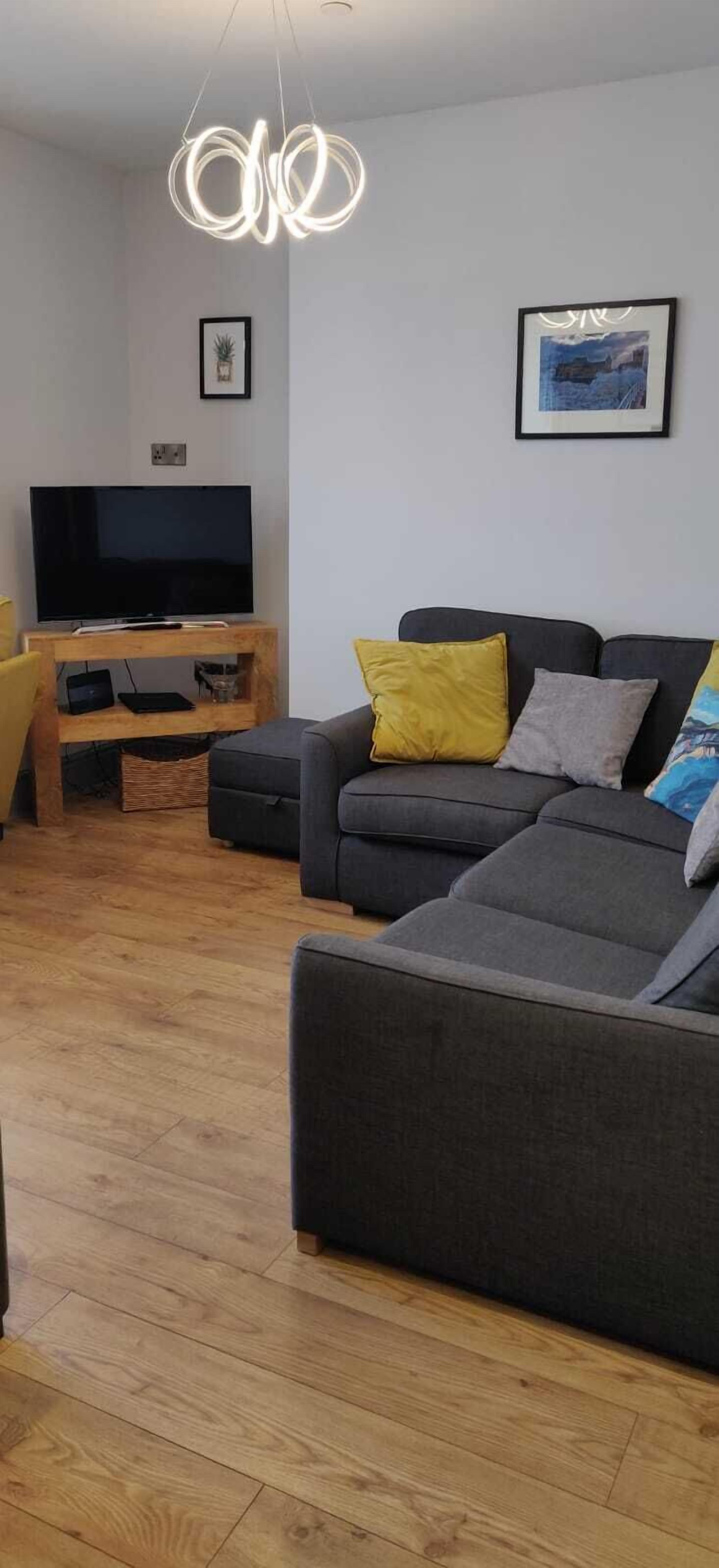 A modern living room with a grey sofa, yellow cushions, a wooden entertainment unit with a TV, and a unique circular light fixture.