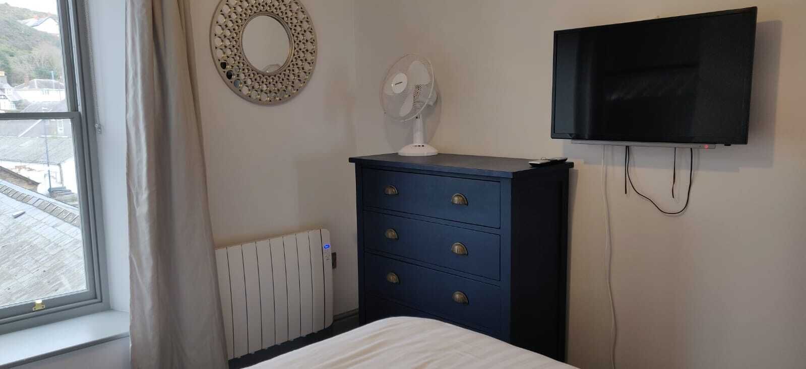 A bedroom with a bed, a dresser, a wall-mounted TV, a radiator, and a window with curtains.