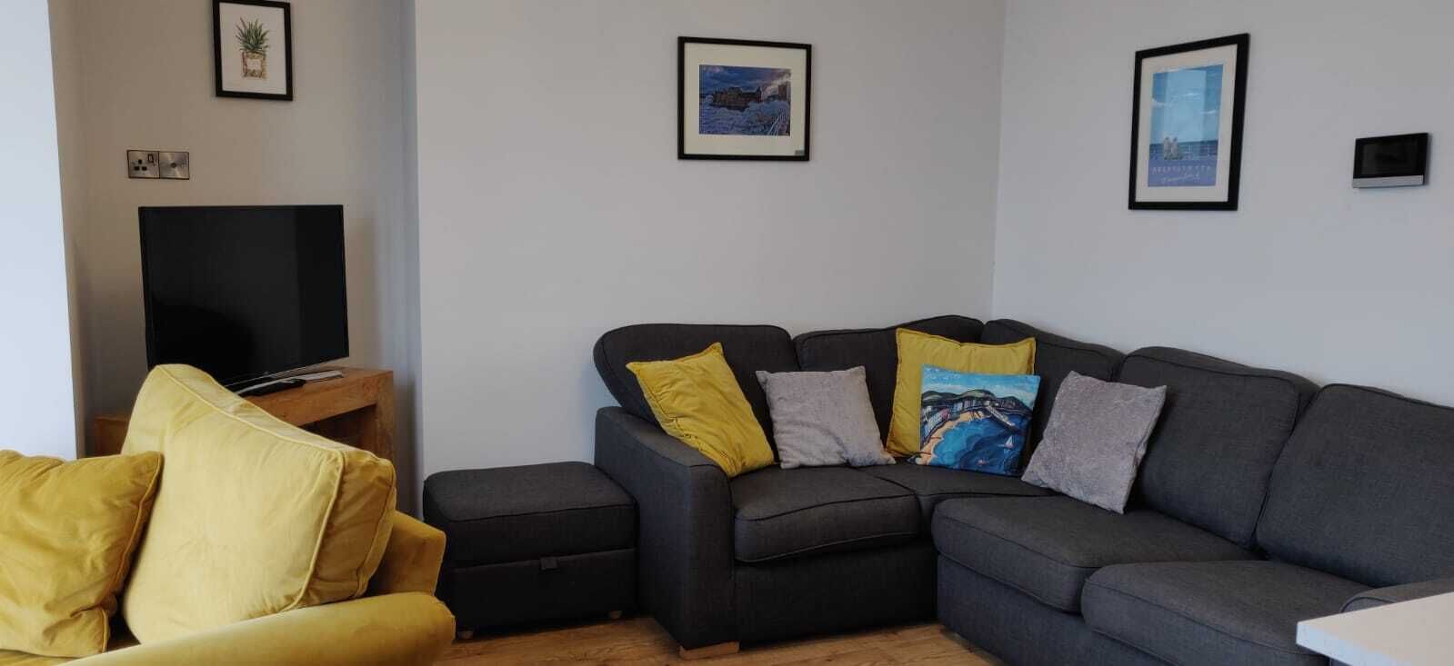 Un salon avec un canapé gris, des coussins jaunes, un petit pouf et un téléviseur à écran plat posé sur un meuble en bois.