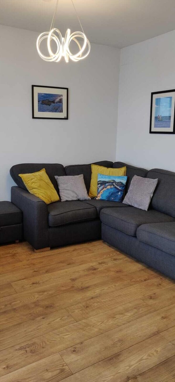 A modern living room with a grey sectional sofa, wooden flooring, and a unique circular light fixture. - Apartment 3 - Belmont  - Aberystwyth (Aberystwyth)