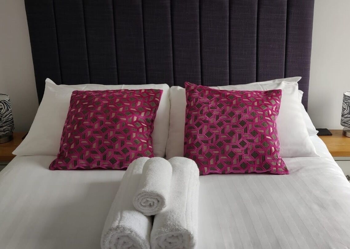 A neatly made bed with white linens, two pink patterned pillows, and two rolled white towels.