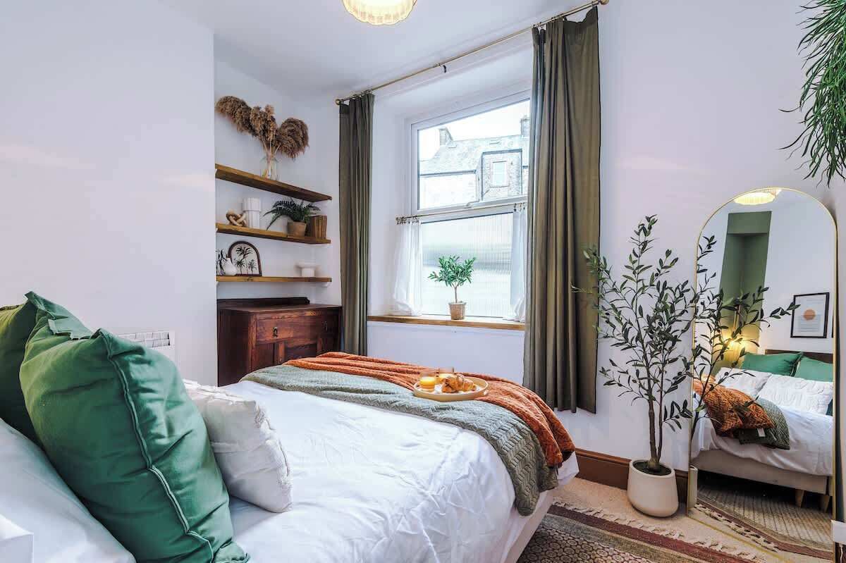 A bedroom with a bed, a window, a mirror, a shelf with decorative items, and a potted plant.
