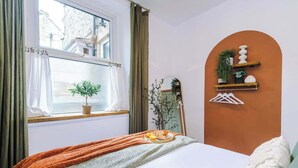 A bedroom with a bed, a window with curtains, a bedside plant, a mirror, and a wall-mounted shelf with decorative items.