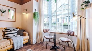 A cozy living room with a leather sofa, a round table, two chairs, a hanging plant, and a window with blinds.