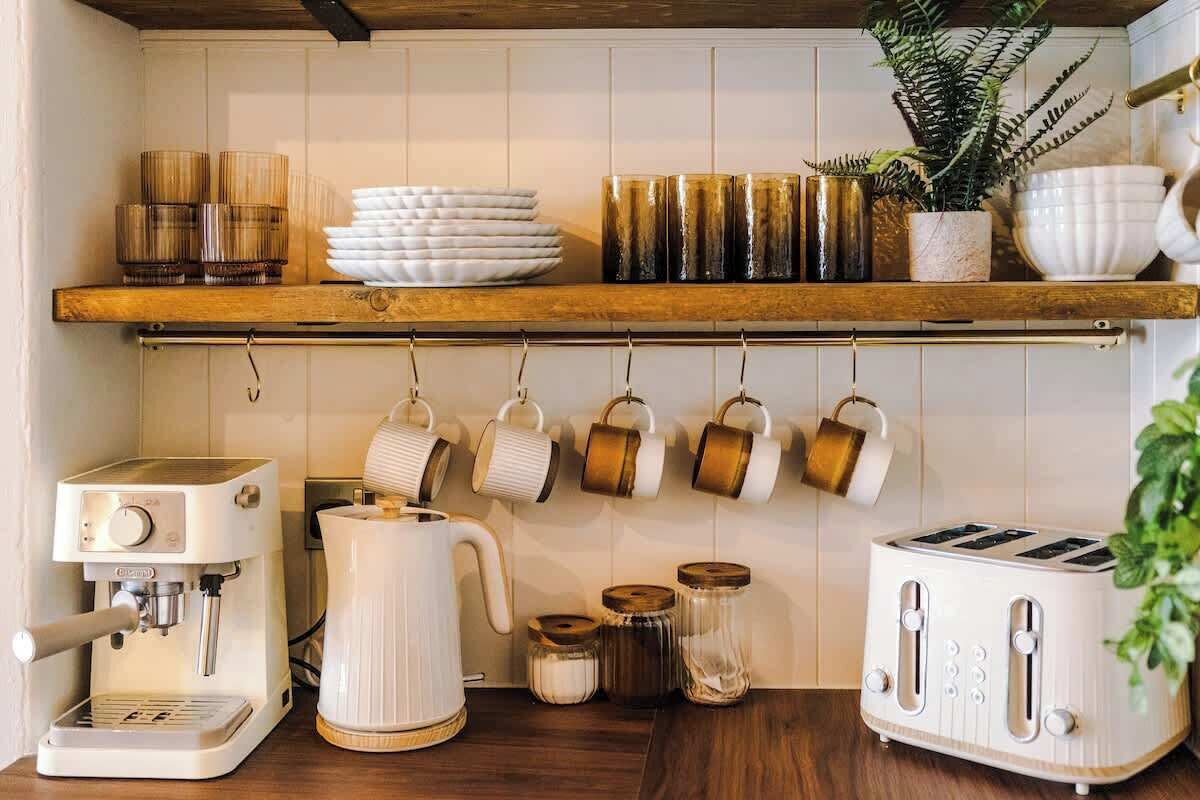 Una cocina con cafetera, tostadora, tazas colgadas y frascos de almacenamiento.