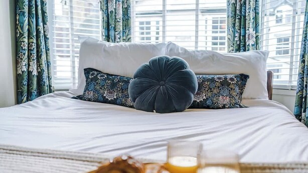 A bed with floral-patterned pillows and a round tufted pillow, a tray with croissants and orange juice, and a window with curtains.