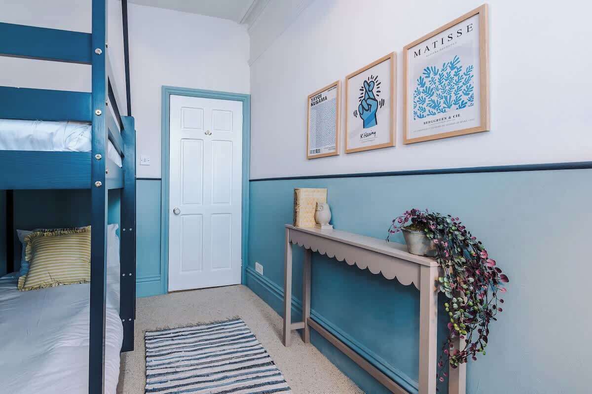 Ein Zimmer mit einem Etagenbett, einer Tür, einem Konsolentisch mit Pflanze und gerahmten Kunstwerken an der Wand.