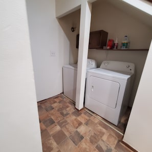 Una sala de lavandería con dos lavadoras, un estante con productos de limpieza y suelo de baldosas.
