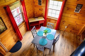 A wooden cabin interior with a dining table, chairs, a small plant, and a window with red curtains.