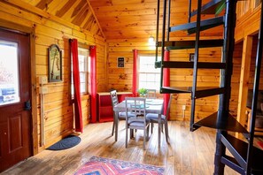 A wooden cabin interior with a dining table, chairs, a bunk bed, and a window with curtains.
