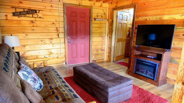 A log cabin interior with a sofa, ottoman, fireplace, TV, and wooden walls. - Welcome to Fisherman's Dream, located near Hocking (McArthur)