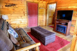 A log cabin interior with a sofa, ottoman, fireplace, TV, and wooden walls.