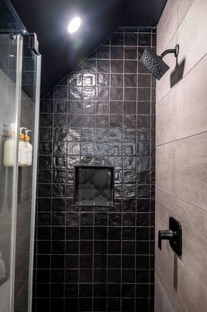 A bathroom with a glass shower enclosure, dark tiled walls, and a mounted showerhead. - Modern Swanky Baseball Home, King Beds, Hot Tub, BBQ Grill, 5 min to Cle Clinic (Cleveland)
