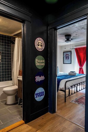 A room with a black door featuring stickers, a bed with a blue blanket, a red curtain, and a framed jersey on the wall. - Modern Swanky Baseball Home, King Beds, Hot Tub, BBQ Grill, 5 min to Cle Clinic (Cleveland)