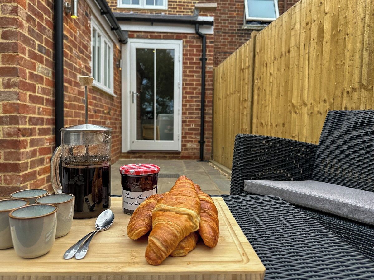 Een terras met een rieten bank, een houten tafel met croissants, een koffiepot en een pot jam.