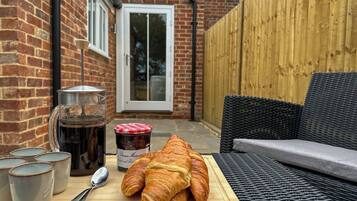 Een terras met een rieten bank, een houten tafel met croissants, een koffiepot en een pot jam.