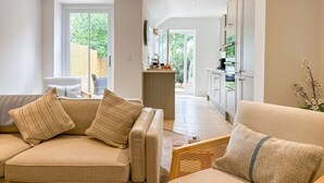 Een moderne woonkamer met een beige bank, een rieten fauteuil en een keuken met witte kasten en een aanrecht.