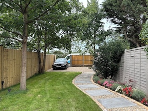 A paved pathway leading to a car parked in a backyard with a wooden fence, a tree, and a flower bed. - Stylish Canterbury Home  • Pet-Friendly, Free Parking (Canterbury)