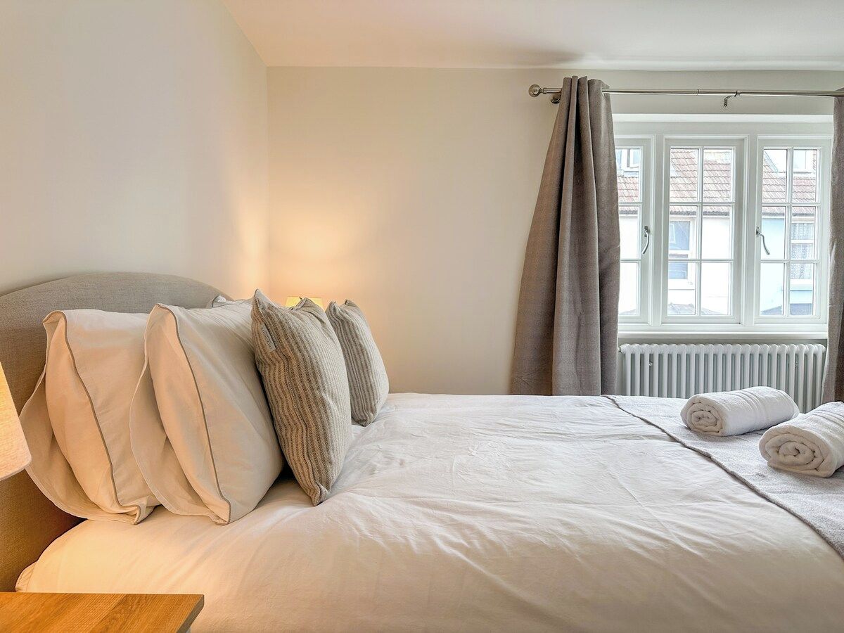 A neatly made bed with pillows and rolled towels, a window with curtains, and a radiator.