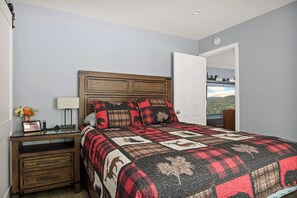 Un dormitorio con cabecera de madera, colcha acolchada con estampados de animales, mesita de noche con lámpara y un florero, y vista a un bosque a través de una puerta abierta.