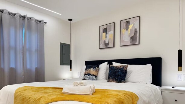 A neatly made bed with a yellow throw blanket, white bedding, and decorative pillows. There are two bedside tables with lamps, a wall-mounted light, and two abstract paintings above the bed.