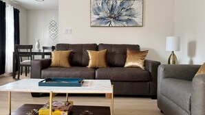 A living room with a brown leather sofa, a coffee table, and a grey armchair. There is a painting on the wall and a lamp on a side table.