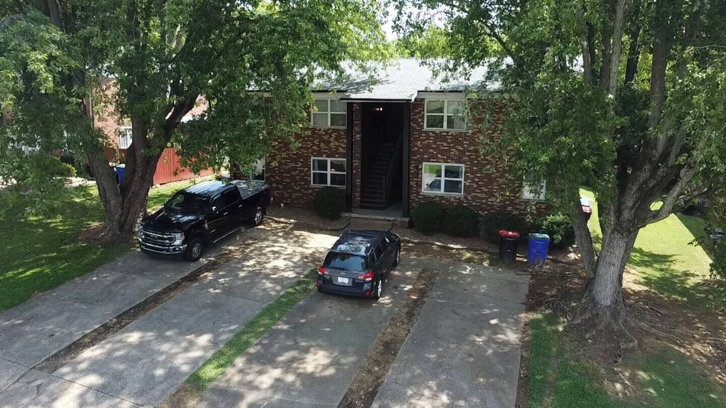 A two-story brick building with a front door, two parked cars, and trees surrounding the property.