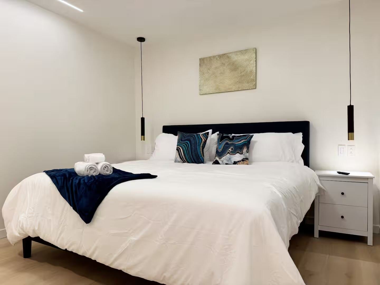 Un dormitorio moderno con una cama grande, ropa de cama blanca, una manta azul oscuro y una almohada decorativa. Hay una mesita de noche con una lámpara y una obra de arte colgada en la pared sobre la cama.