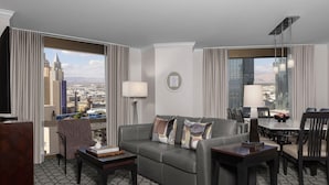 Une chambre d’hôtel moderne avec un canapé en cuir gris, une petite table basse, un coin repas avec une table et des chaises, et de grandes fenêtres offrant une vue sur la ville.