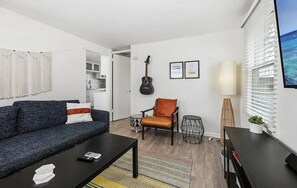 A living room with a sofa, a chair, a coffee table, a guitar, framed pictures, and a window with blinds. - A Charming Hidden Gem, two blocks to Moonlight Beach (Encinitas)