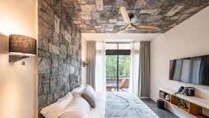 A bedroom with a stone wall, a ceiling fan, a bed with white linens, a TV on the wall, and a balcony with a view of greenery. - Hotel-Style King Room • Private Terrace with Jetted Dip Pool (Tulum)